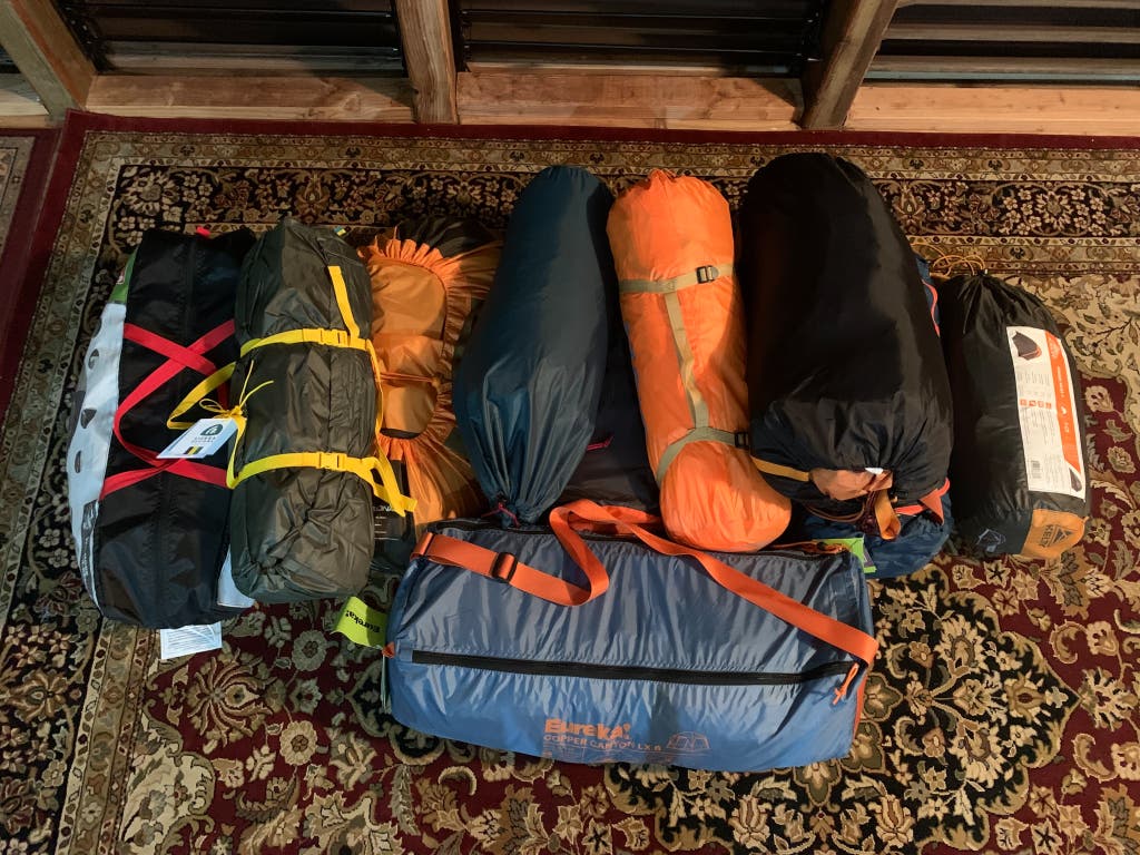 A variety of camping tents packed inside their colored carrying bags laid out on a floor for comparison.
