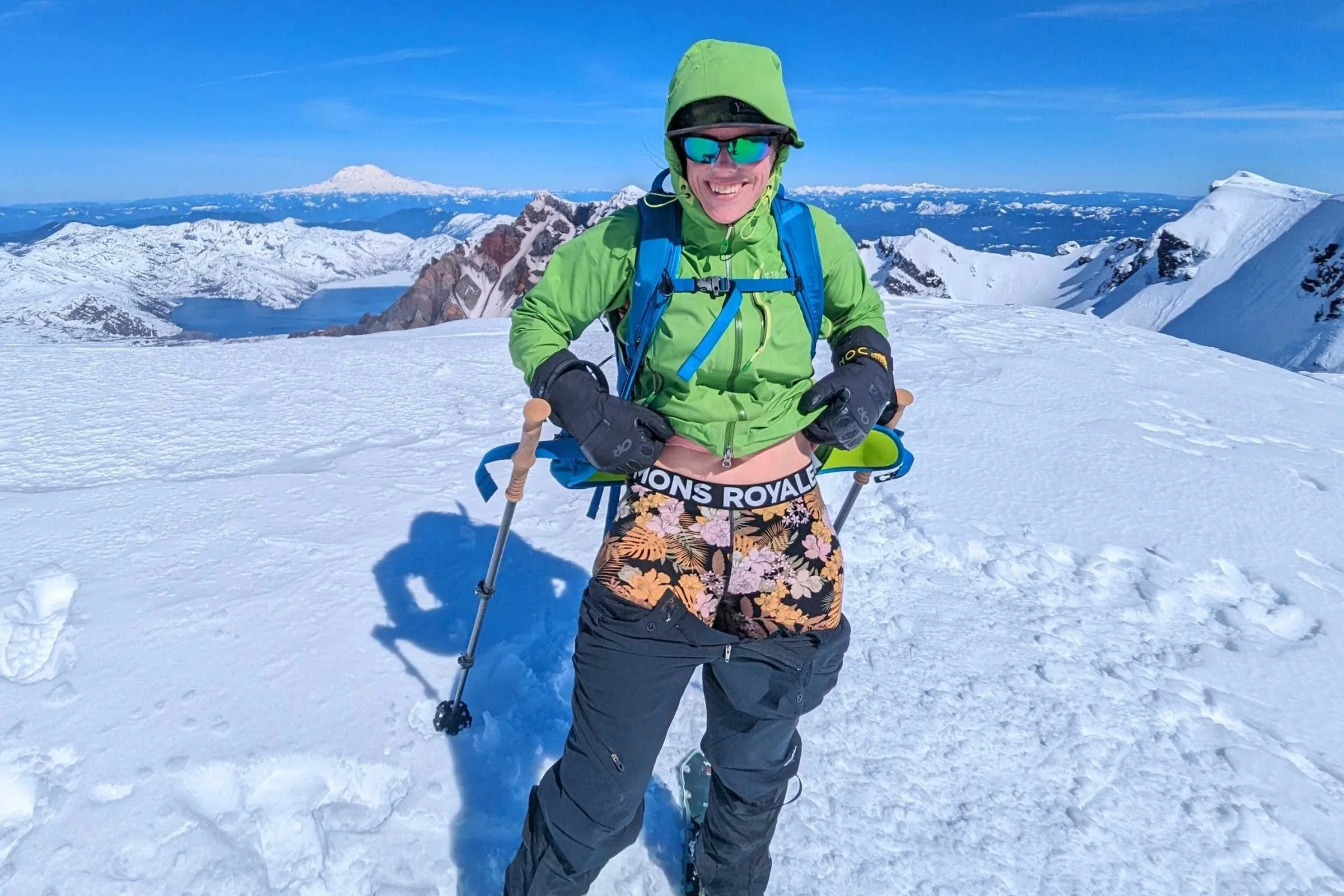 A woman standing triumphantly on a snowy mountain summit wearing patterned leggings.