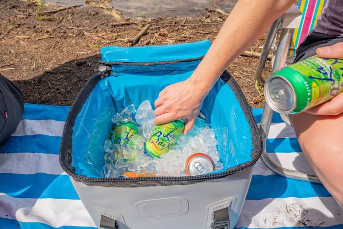 A close-up of a hand placing beverage cans into a soft cooler filled with ice.