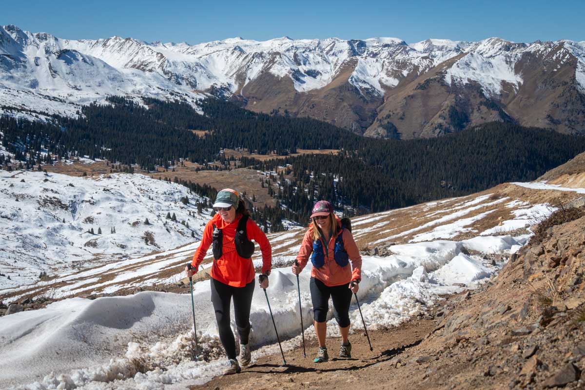 Two runners using trekking poles to navigate a steep, snow-covered mountain ridge.