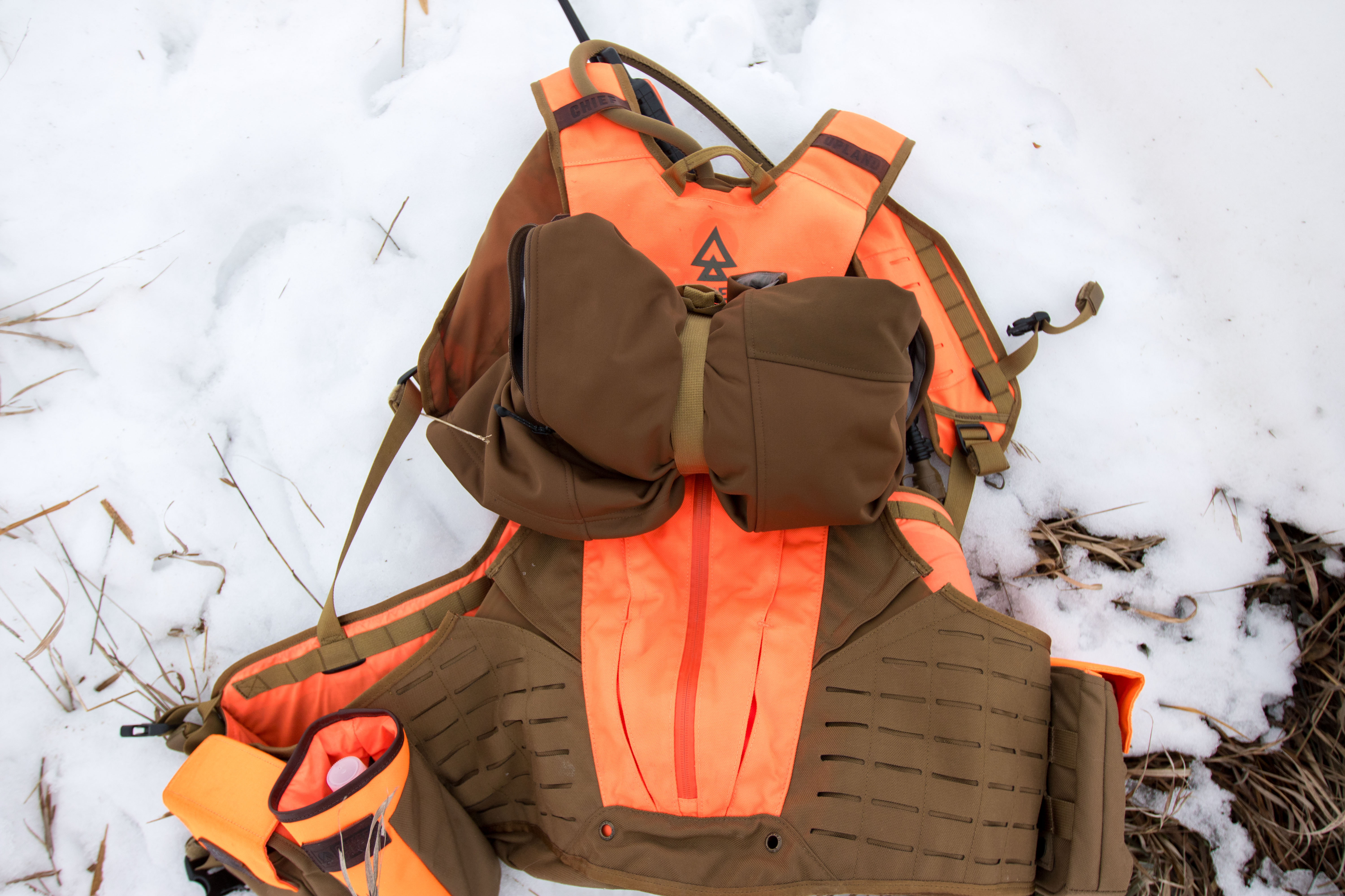 A hunting vest lying in the snow with an orange jacket strapped to the exterior.