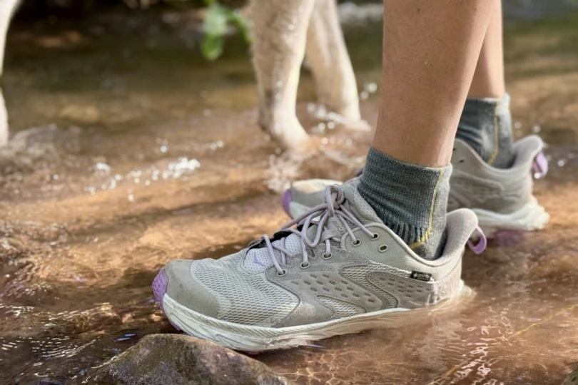 A hiker wearing Hoka Anacapa Low GTX shoes stepping through a shallow mountain stream.