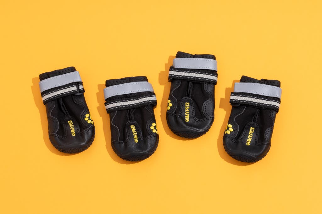 Four black Qumy Dog Shoes with velcro straps on a yellow background.