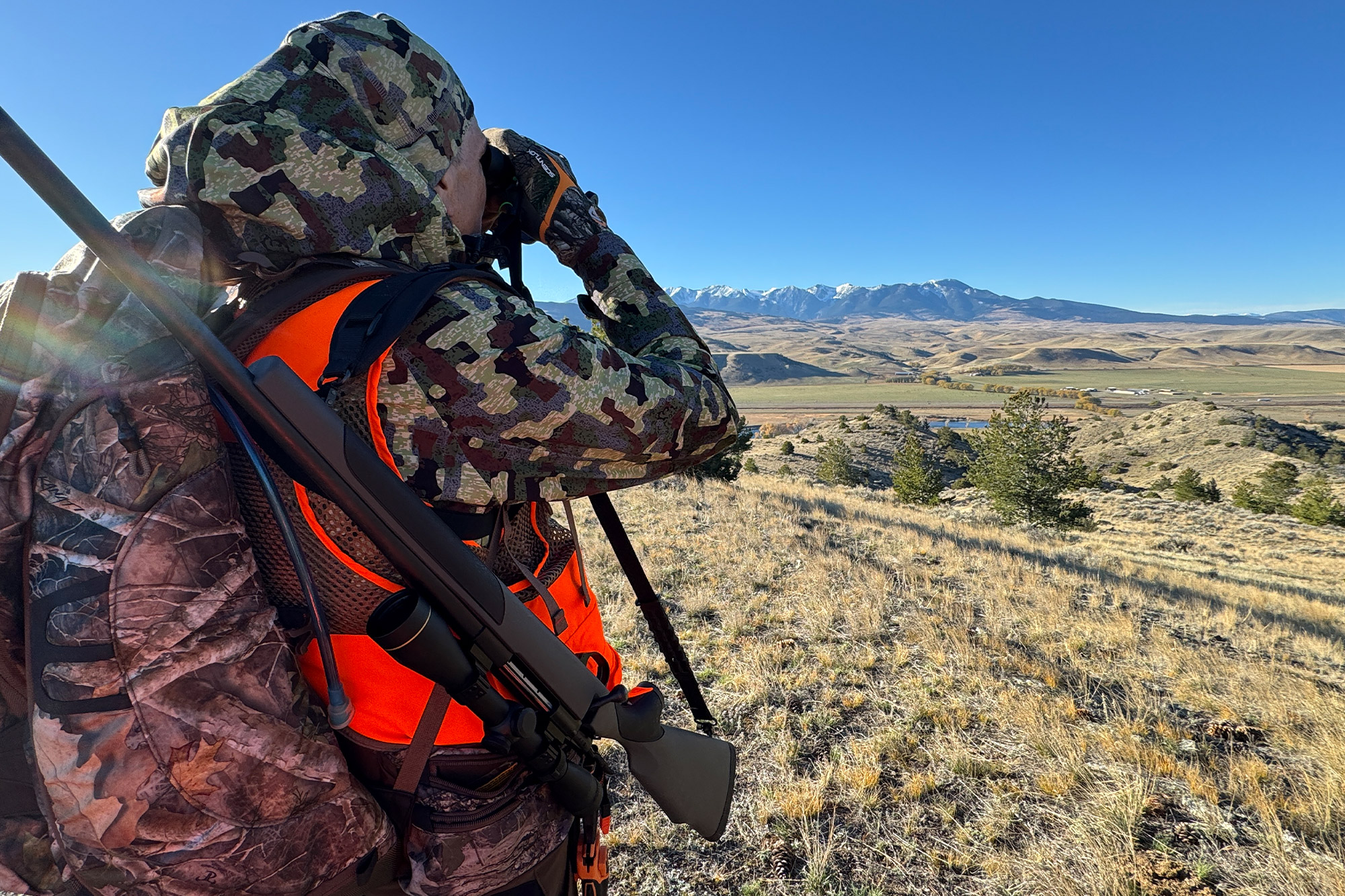 Close-up of the FORLOH camo pattern on a jacket while the hunter uses binoculars on a grassy slope.