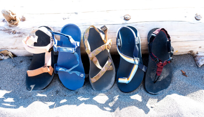 A lineup of various hiking sandals on a sandy beach.