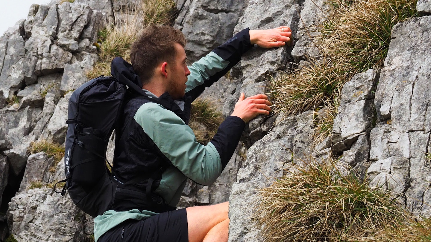 A hiker wearing technical shorts and a backpack scrambling up a steep rock face.