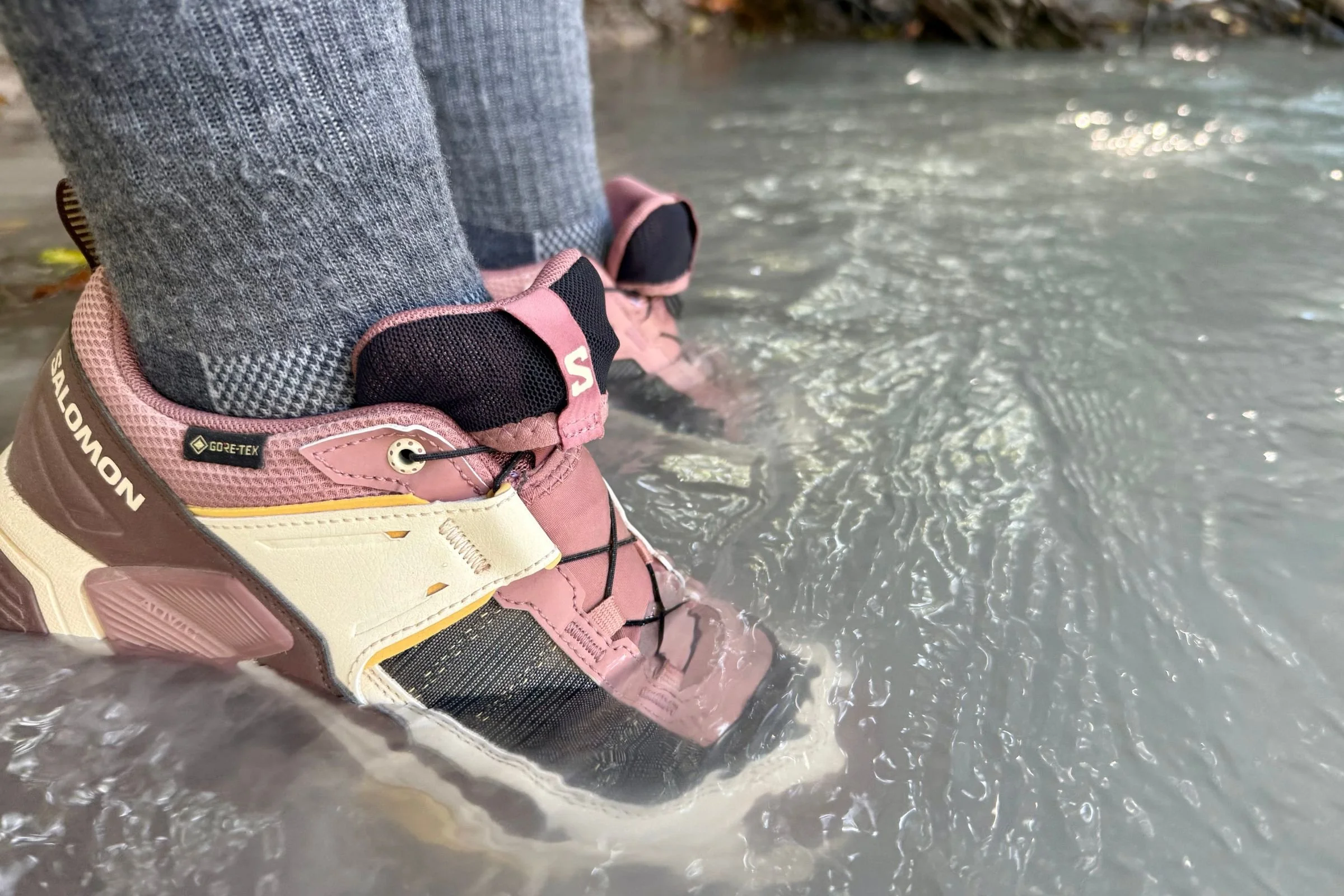 A Salomon hiking shoe being submerged in a shallow river to test its Gore-Tex waterproofing.
