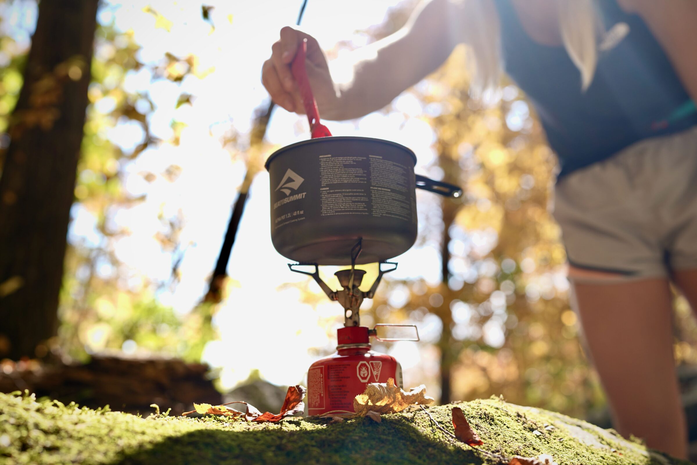 Hands stirring a small camping pot on a portable stove in the wilderness.