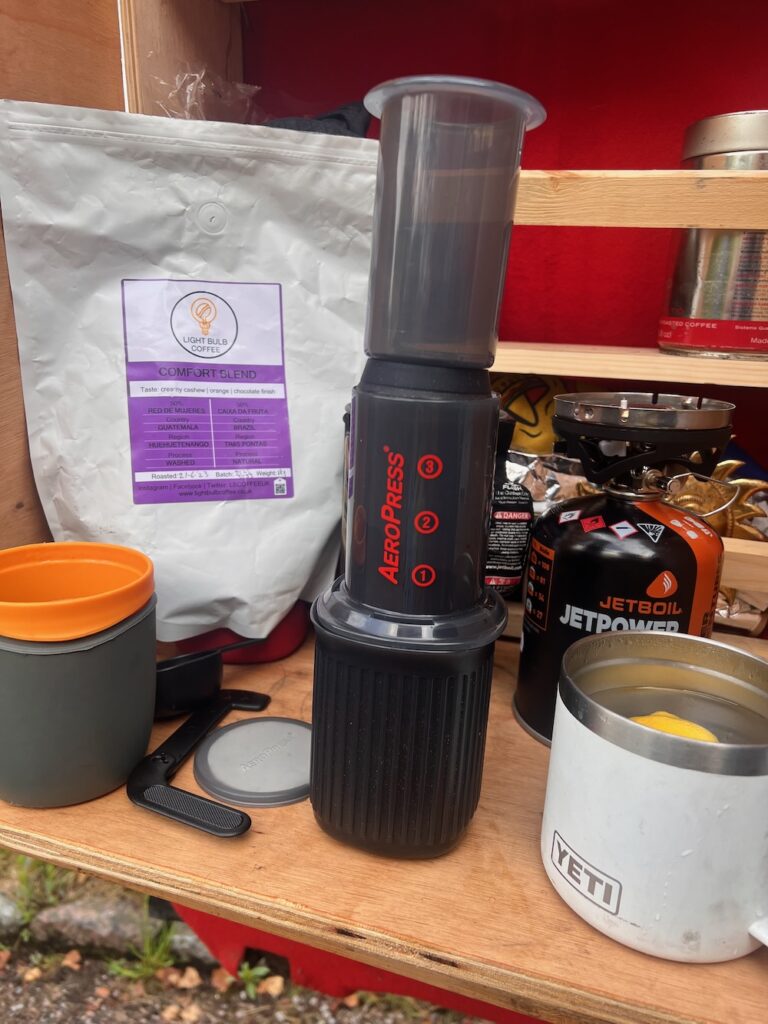 A person using an AeroPress coffee maker at a campsite to brew coffee manually.